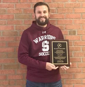 Coach Lewis poses with his plague in front of a brick wall.