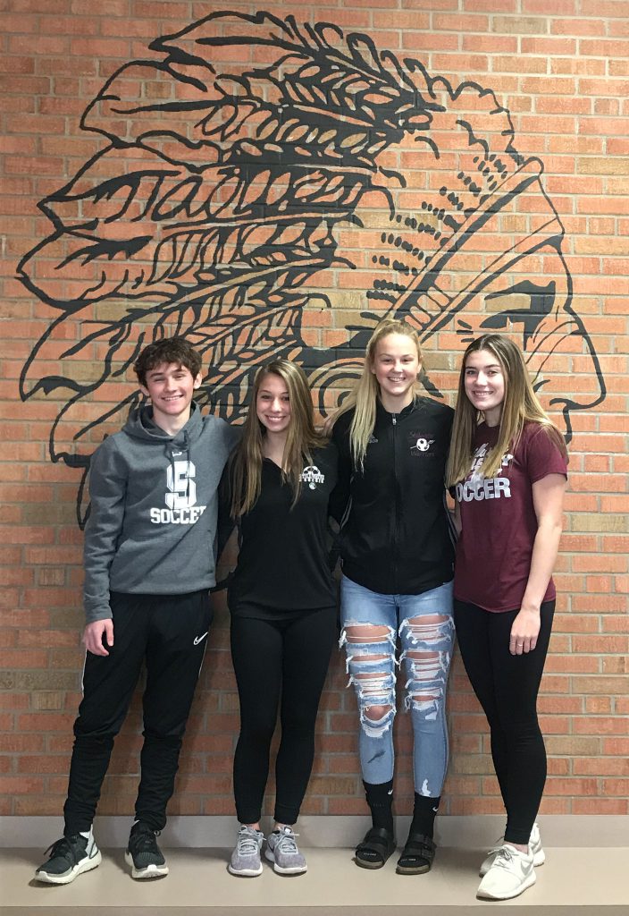 Four students standing in front of warrior head on a brick wall