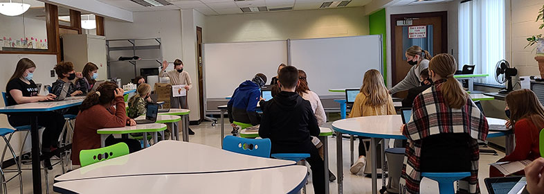 student at a podium holding his hand up while students are seated and standing in the makerspace room, conducting a mock trial