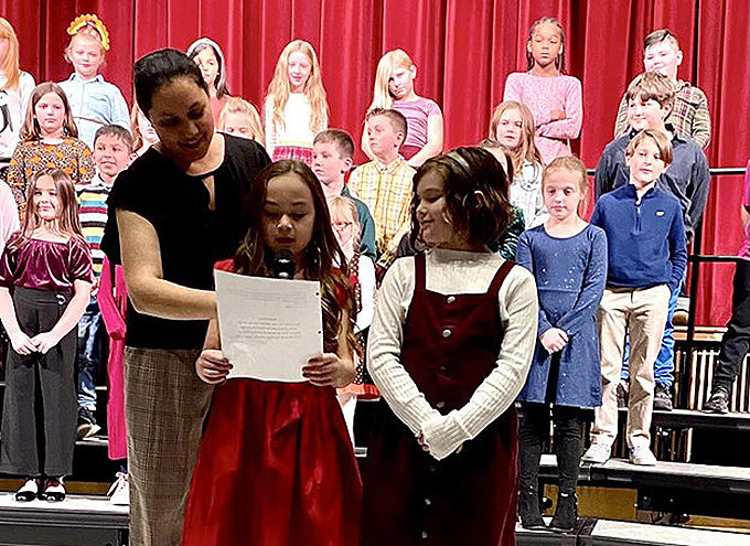 teacher with two students and a group of students behind them on stage