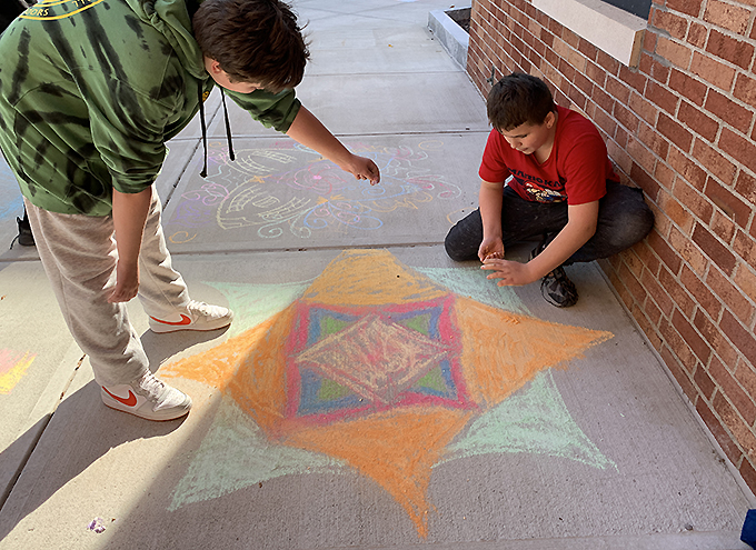 2 students creating a rangoli in front of school using chalk
