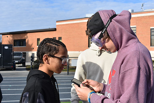 student wearing goggles typing into cell phone while other students look on