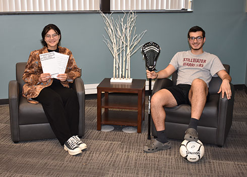 Nurshinta,holding sheet music, and Amoun,holding a lacrosse stick with foot on a soccer ball, seated in chairs