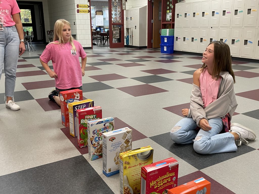 Two students sitting on the floor with cereal boxes in front of them.