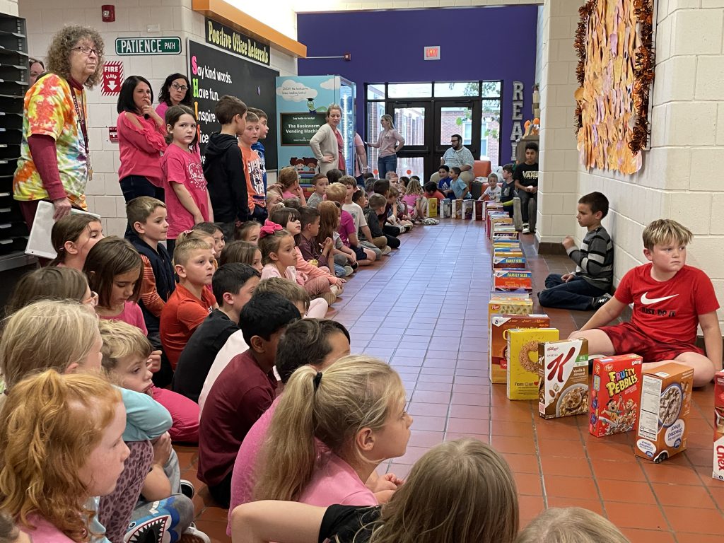 Elementary students sitting on the side of the school hallways with cereal boxes lined up in front of them.