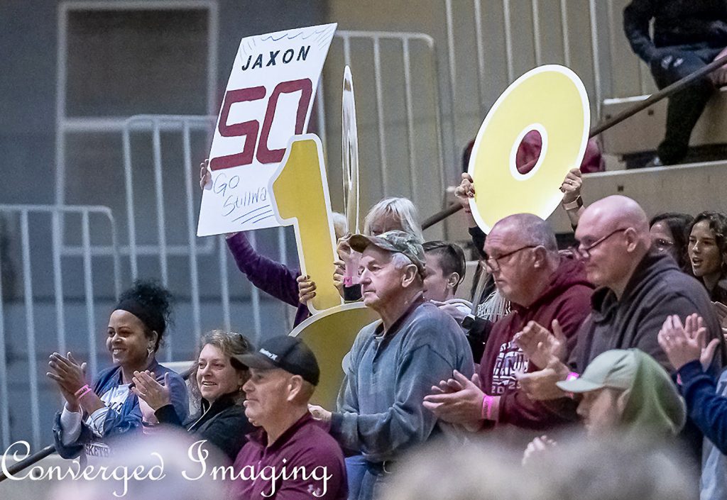 Families and spectators sitting in the stands. One fan is holding a "Jaxon 50" sign.