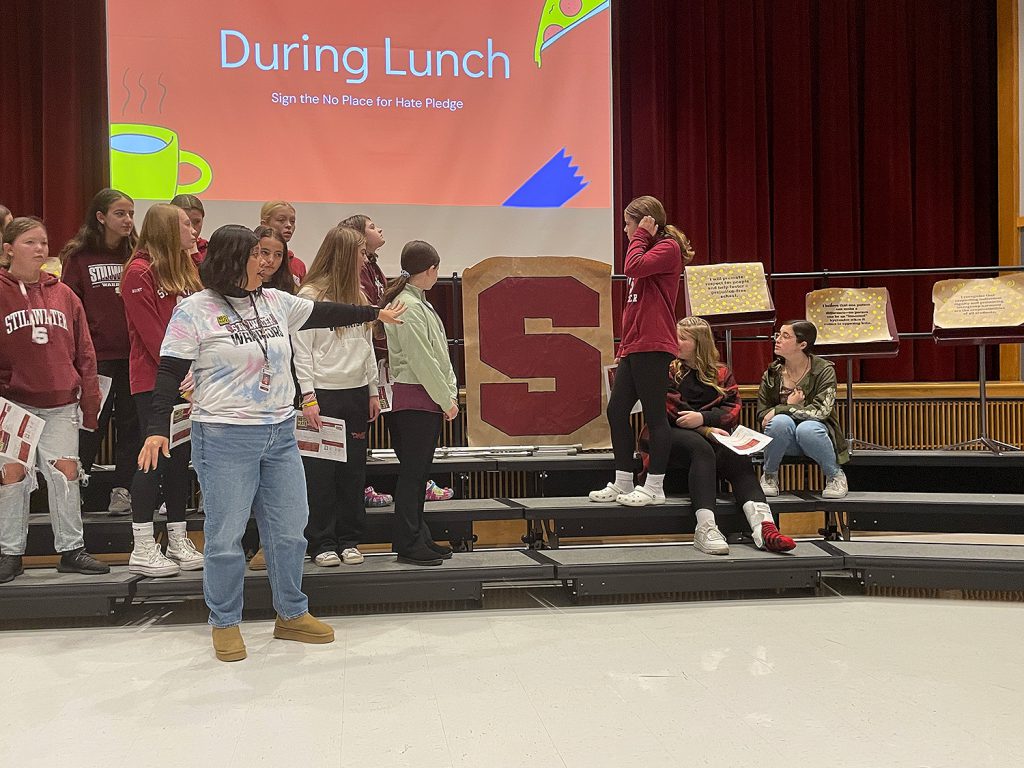 Teacher standing with students on stage pointing to giant "S" drawn on paper.