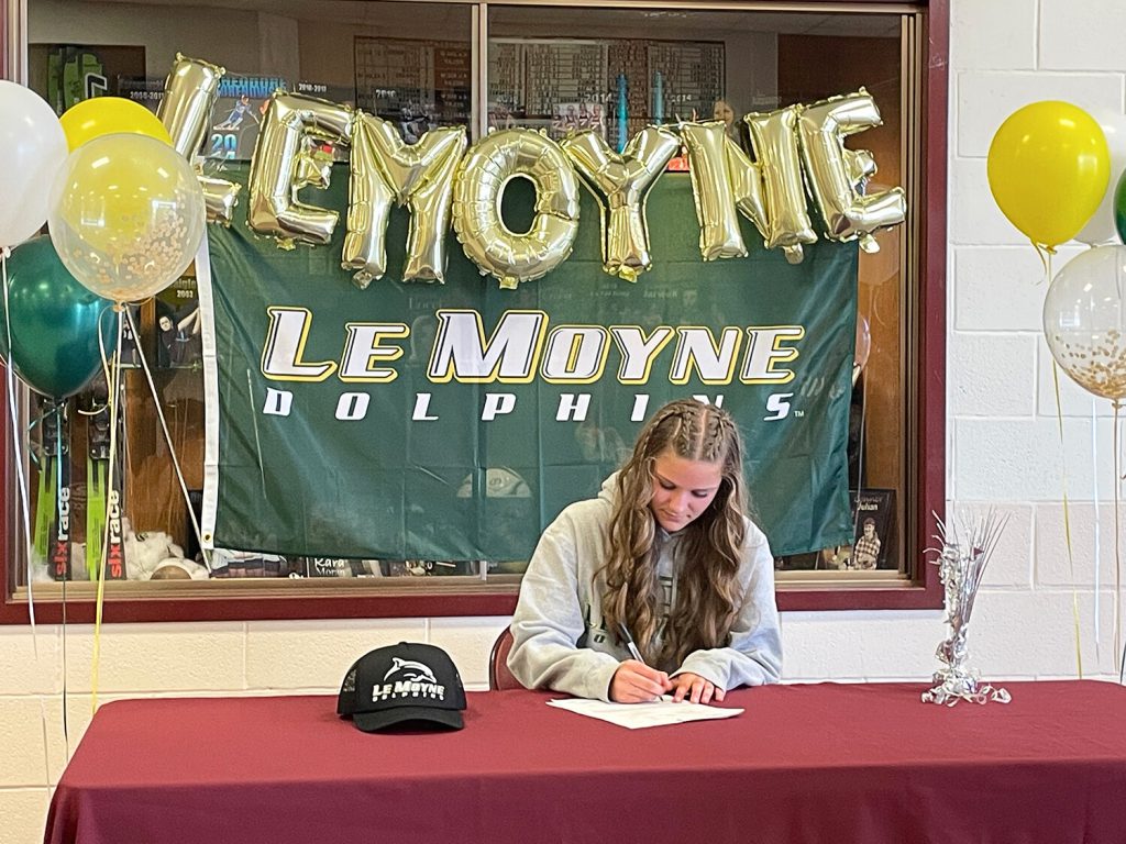 Student signing a letter on a table with "LeMoyne" spelled in gold balloons in the background.