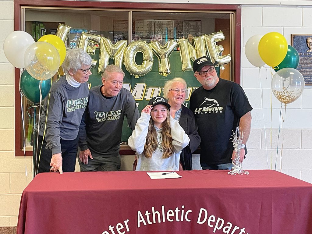 Student with her grandparents smiling for a photo as she wears a black baseball cap with the name of her selected college on it.