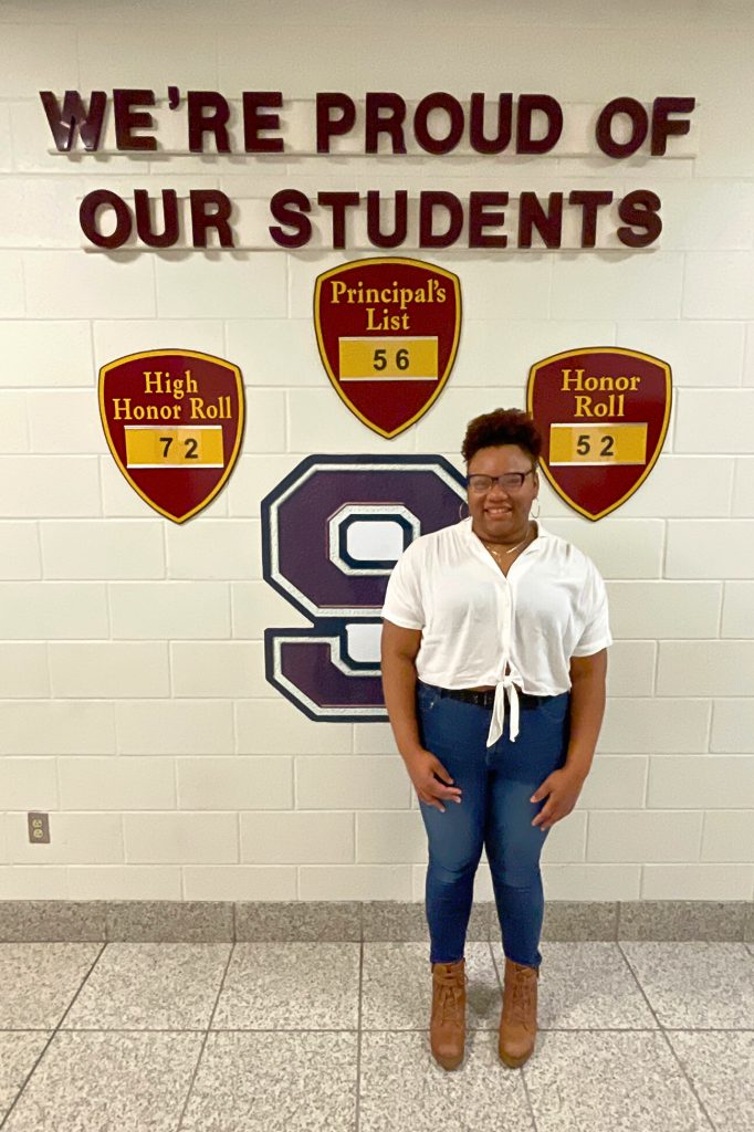 Student smiling in front of wall that says "We're Proud of our Students."