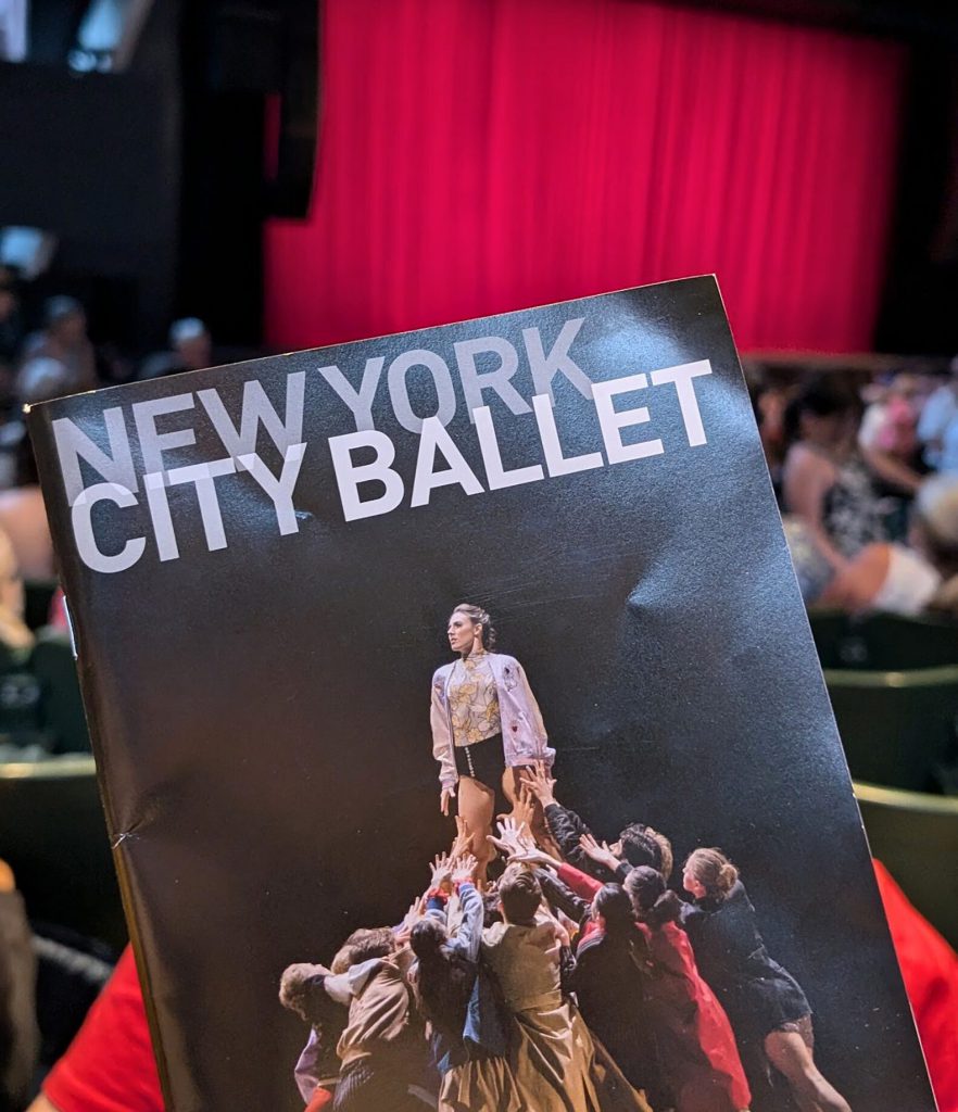 Photo of a New York City Ballet booklet. One person in middle surrounded by group of people reaching out with their hands.