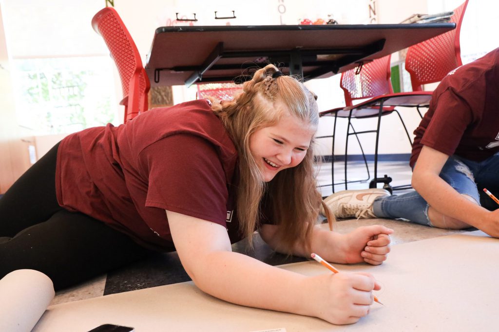 A student happily giggling with a pencil in hand on the ground.