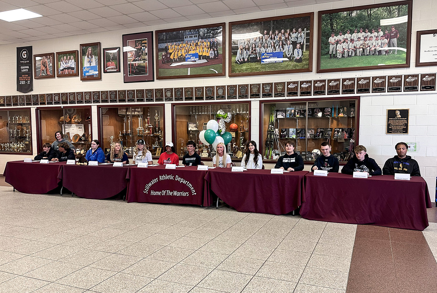 Group of athletes smiling and seated at a table eagerly awaiting letters to sign.