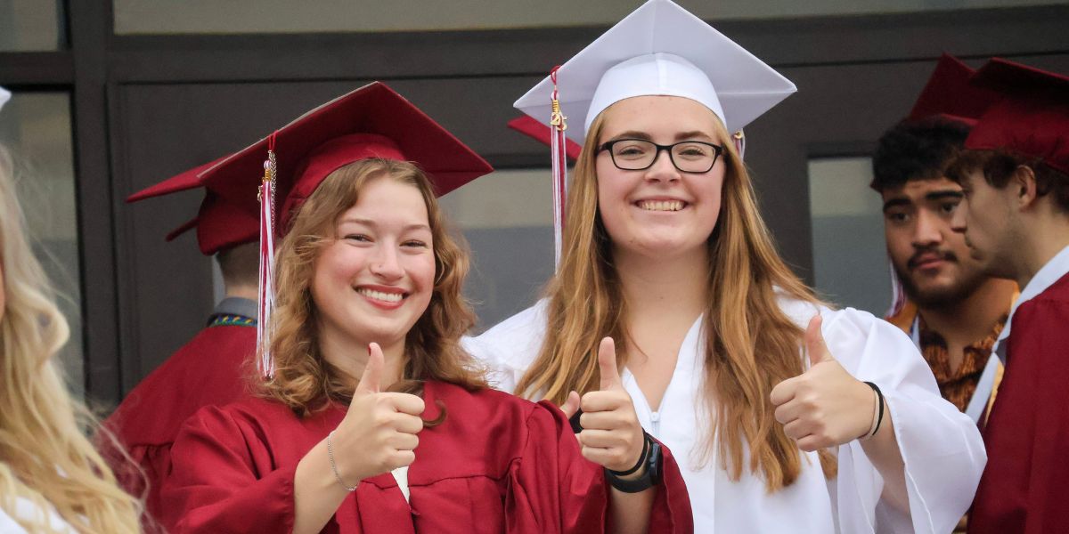 Two graduates smiling and giving a thumbs up to the camera. Graduate of the left is dressed in a maroon cap and gown and the graduate on the right is wearing a white cap and gown.