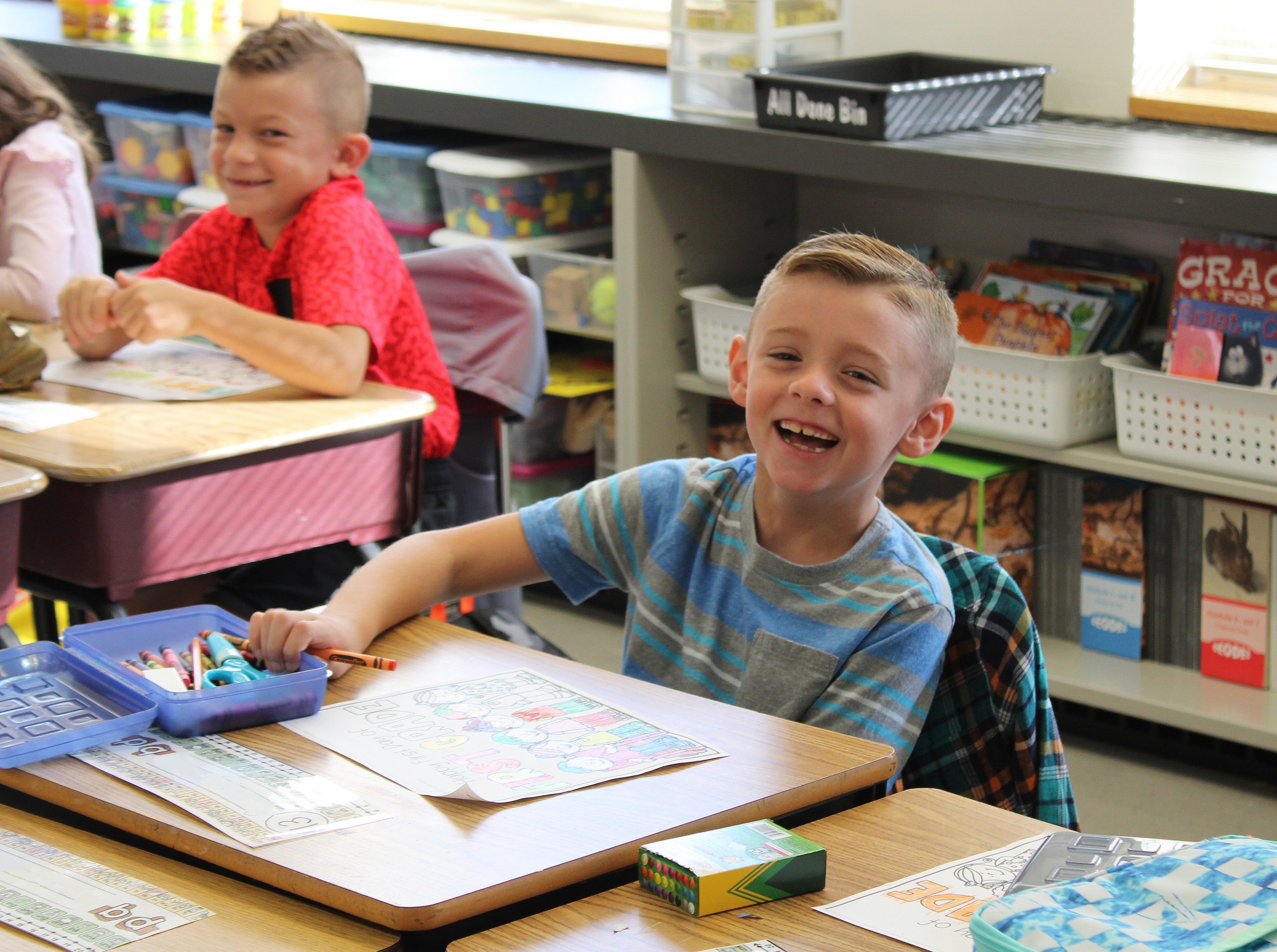 Elementary student smiling while sitting at his desk with a crayon in his hand a drawing in front of him.