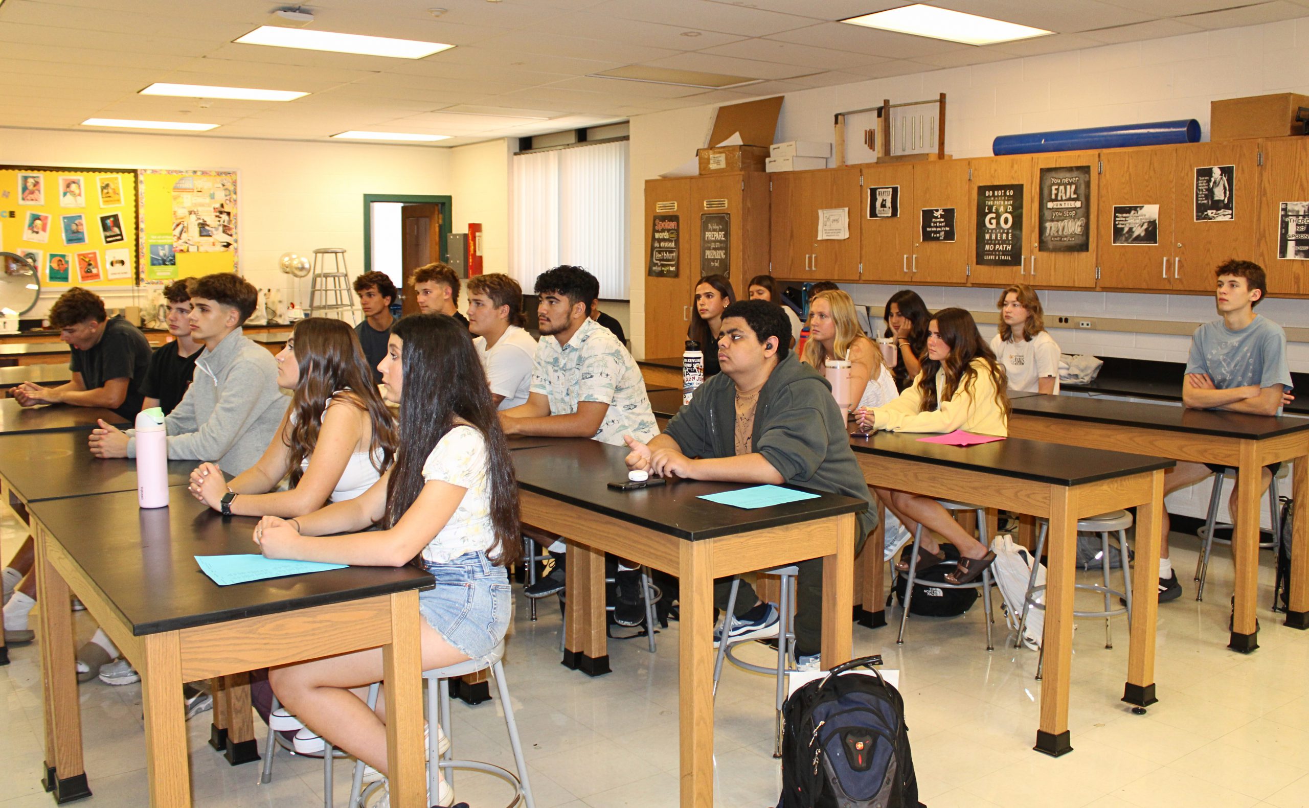 High school students sitting attentively at lab tables in a classroom, facing forward during a lesson or presentation.