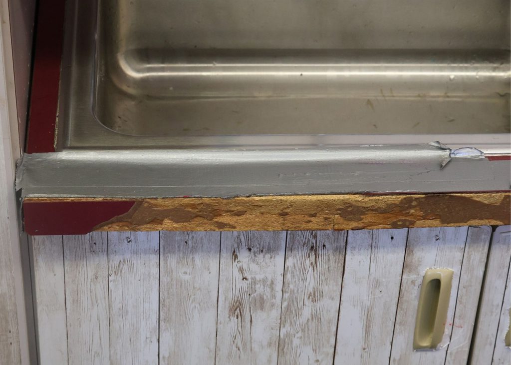 Up close photo of a metal sink within a countertop. The front part of the countertop is being held together with a piece of duct tape. The edge in front is discolored. A small portion on the left side is maroon while the remaining is cracked wood.