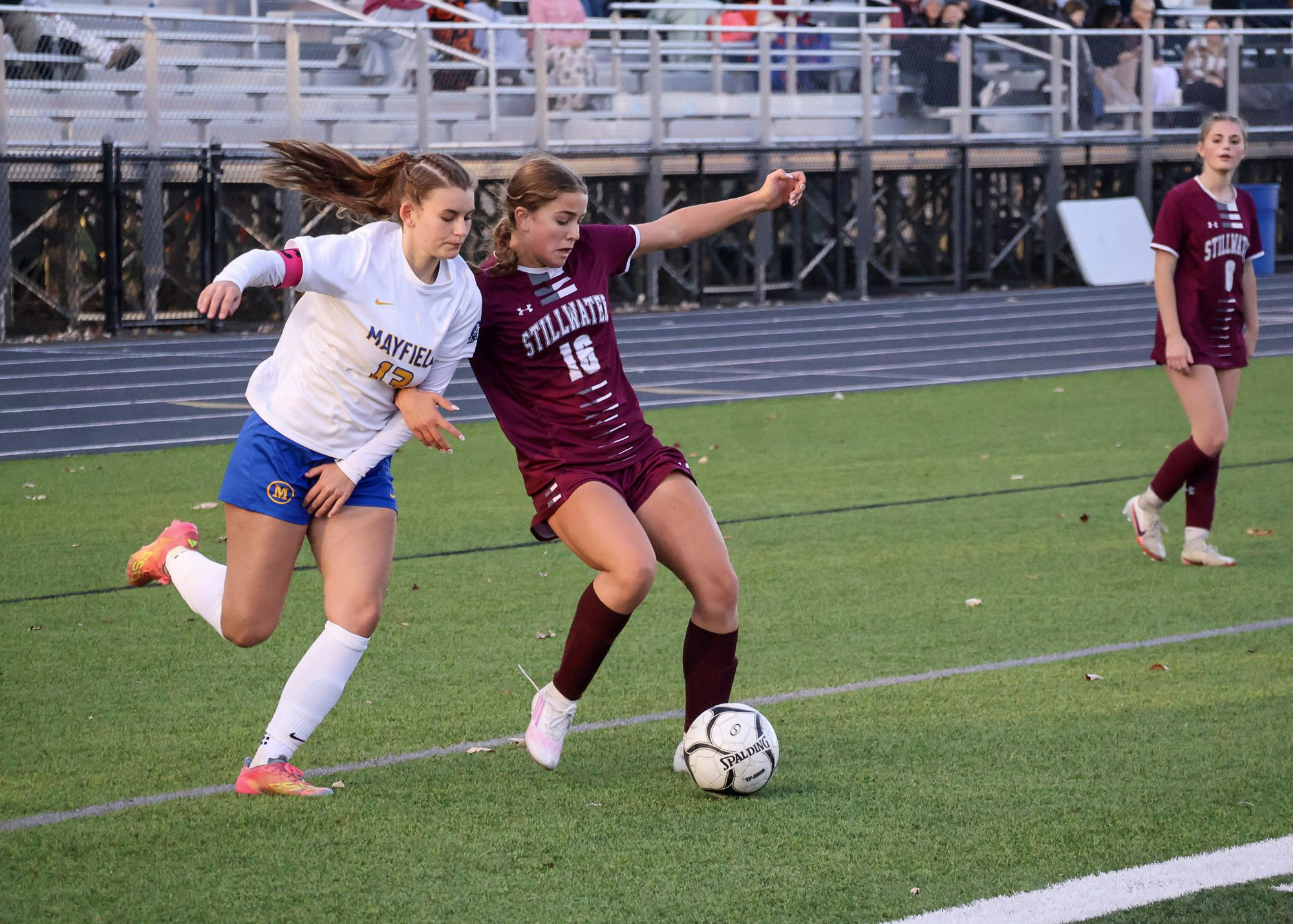 Two high school girl soccer players from opposite teams competing for the ball on the turf field. One girl is wearing a white jersey with blue shorts and the other player is wearing a maroon jersey and shorts. The girl in the maroon jersey is fending off the other player as she attempts to kick the ball.