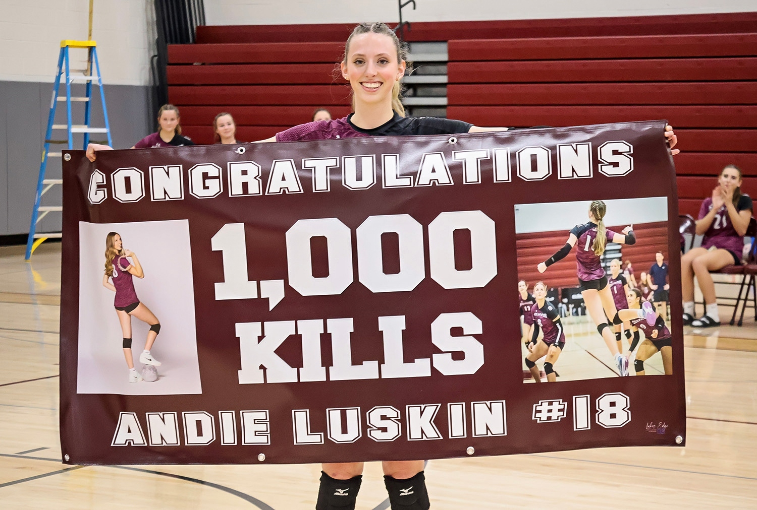 Stillwater High School volleyball player Andie Luskin smiles while holding a large maroon banner that reads “Congratulations 1,000 Kills Andie Luskin #18.” The banner includes two photos of her playing volleyball. Teammates and bleachers are visible in the background of the gym.