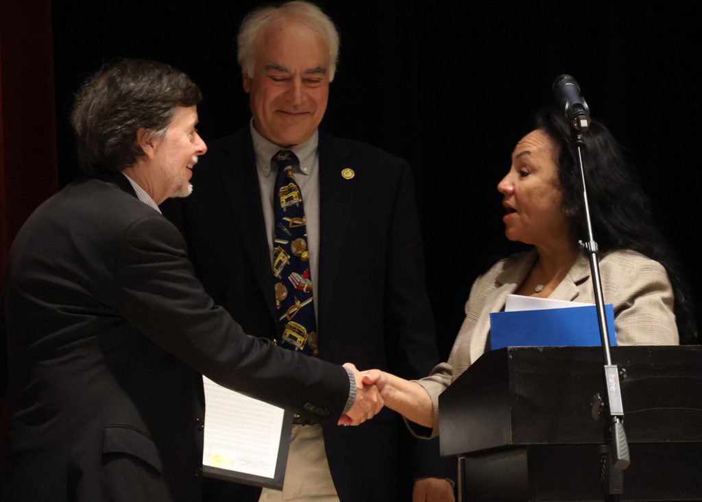A man and a woman shaking hands as the woman presents an award to the man. Another man stands smiling behind them. There is also a podium with a microphone in front of the woman.