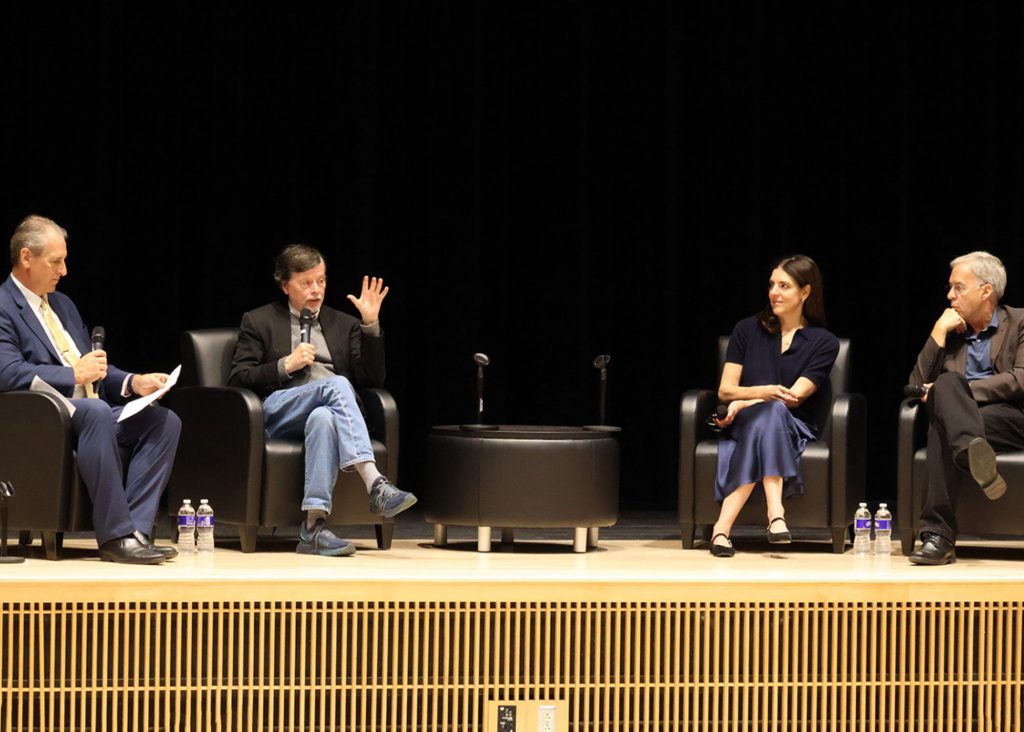 Group of four people sitting on black leather chairs on stage, engaged in a discussion. Each is holding a microphone in their hand.