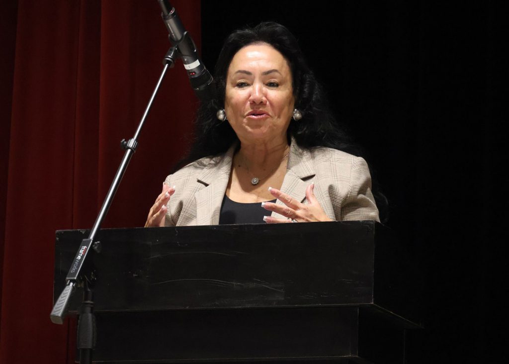 Woman standing at a podium speaking at a microphone. She is gesturing with both of her hands.