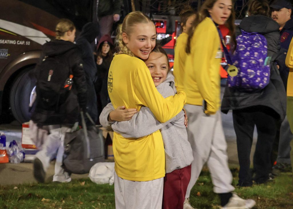 A female soccer player wearing a long sleeve yellow shirt hugging her younger sibling. Both are smiling.