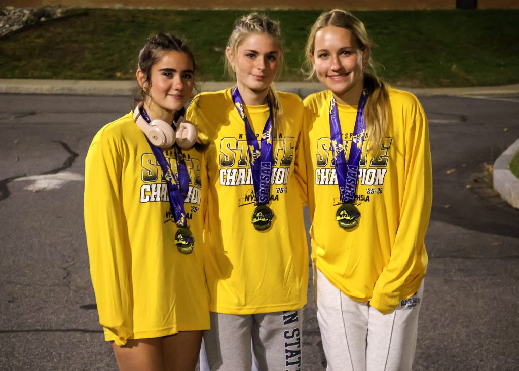 Three female soccer players wearing long sleeve yellow shirts and medals around their necks, smiling for a photo. They are outside at night.