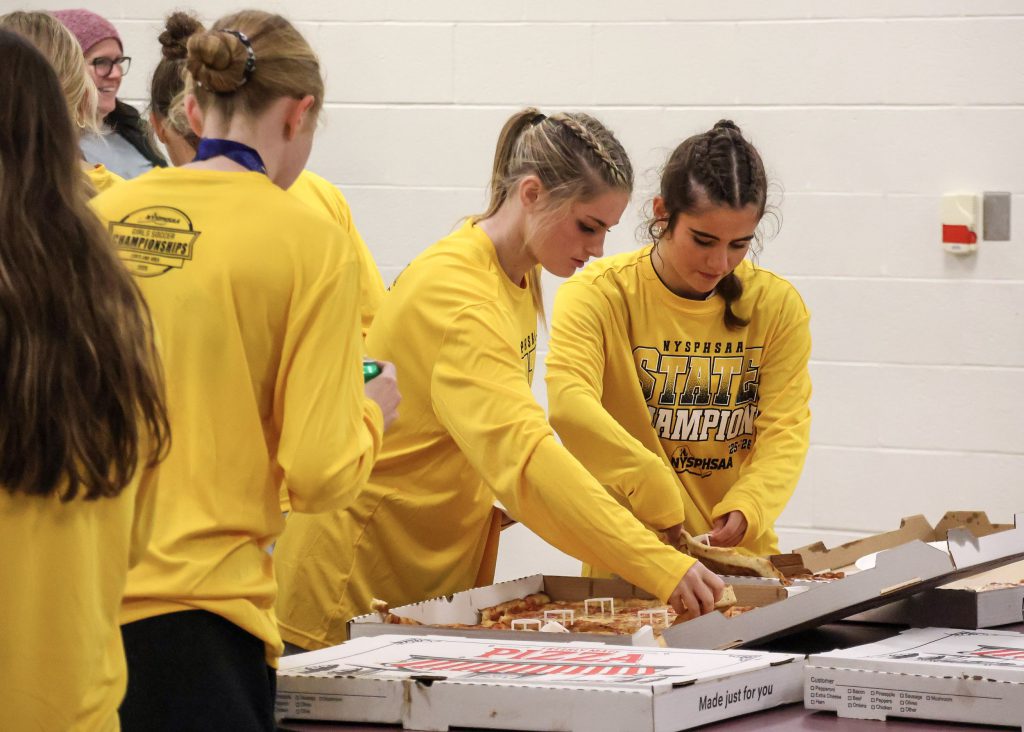 Female soccer players wearing yellow long sleeve shirts grabbing slices of pizza.