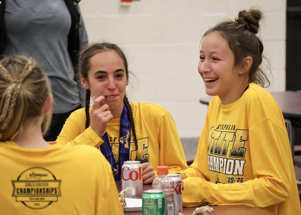 Two female soccer players wearing long sleeve yellow shirts laughing and smiling at a table.