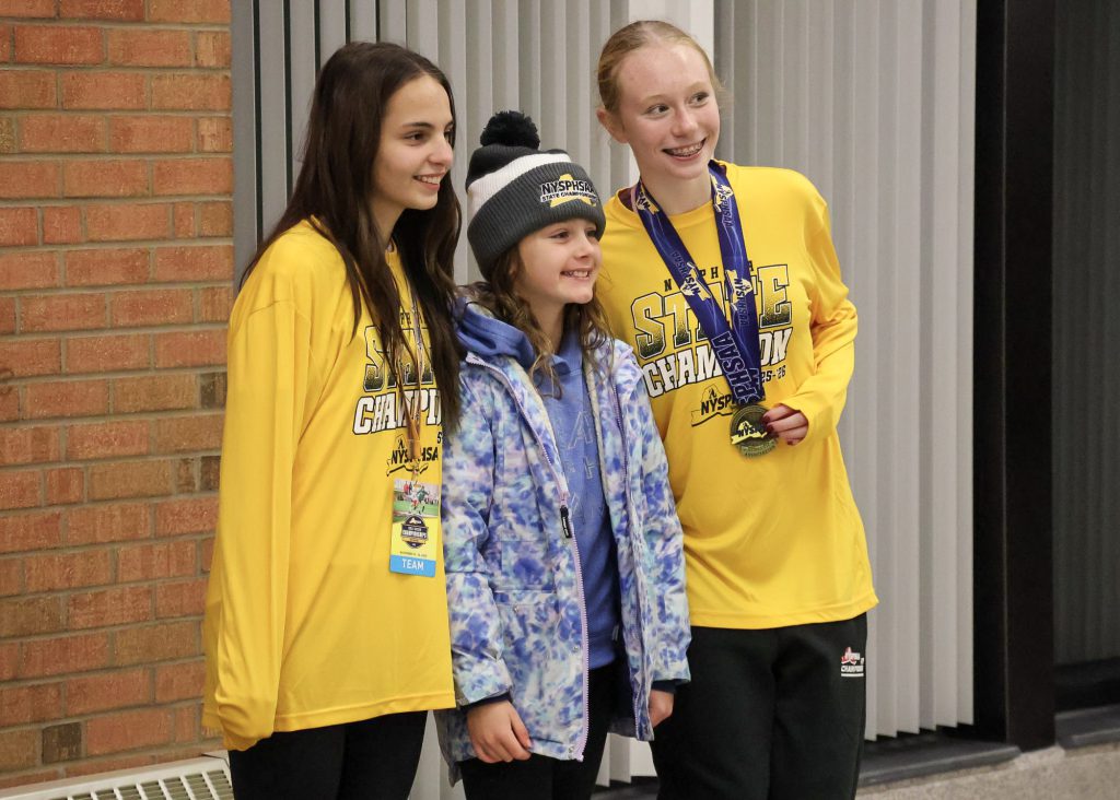 Two female soccer players wearing long sleeve yellow shirts standing with a young girl, who's wearing a blue printed jacket and gray/white striped pom pom beanie that says "NYSPHSAA" on it. The player on the right is wearing a medal around her neck. The three of them are smiling for a photo.