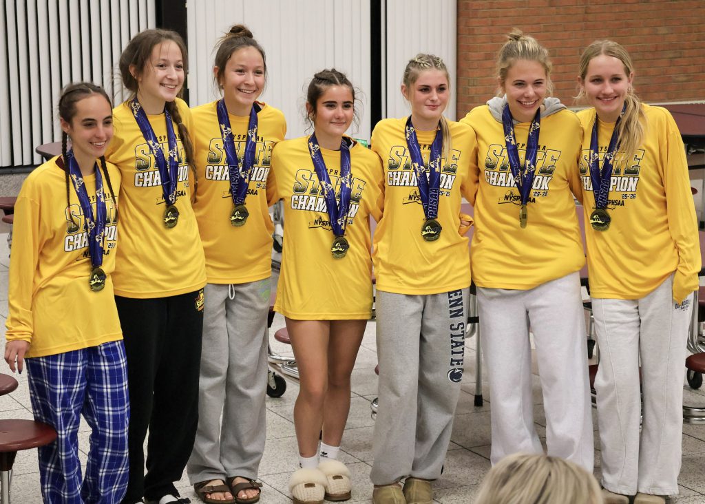 Seven female soccer players wearing long sleeve yellow shirts and medals around their necks, smiling for a group photo.