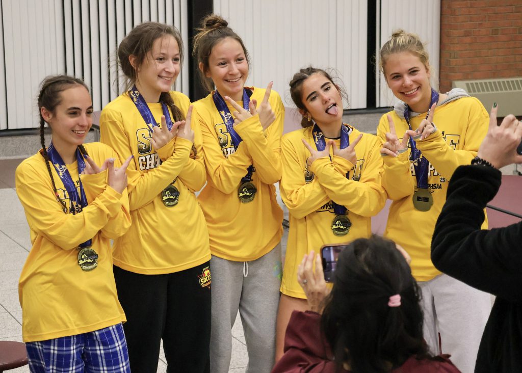 Five female soccer players wearing yellow long sleeve shirts and medals around their necks. They are making a "W" symbol with their hands and posing for a photo.