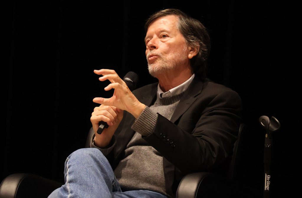 A man speaking into a microphone as he gestures with his right hand.