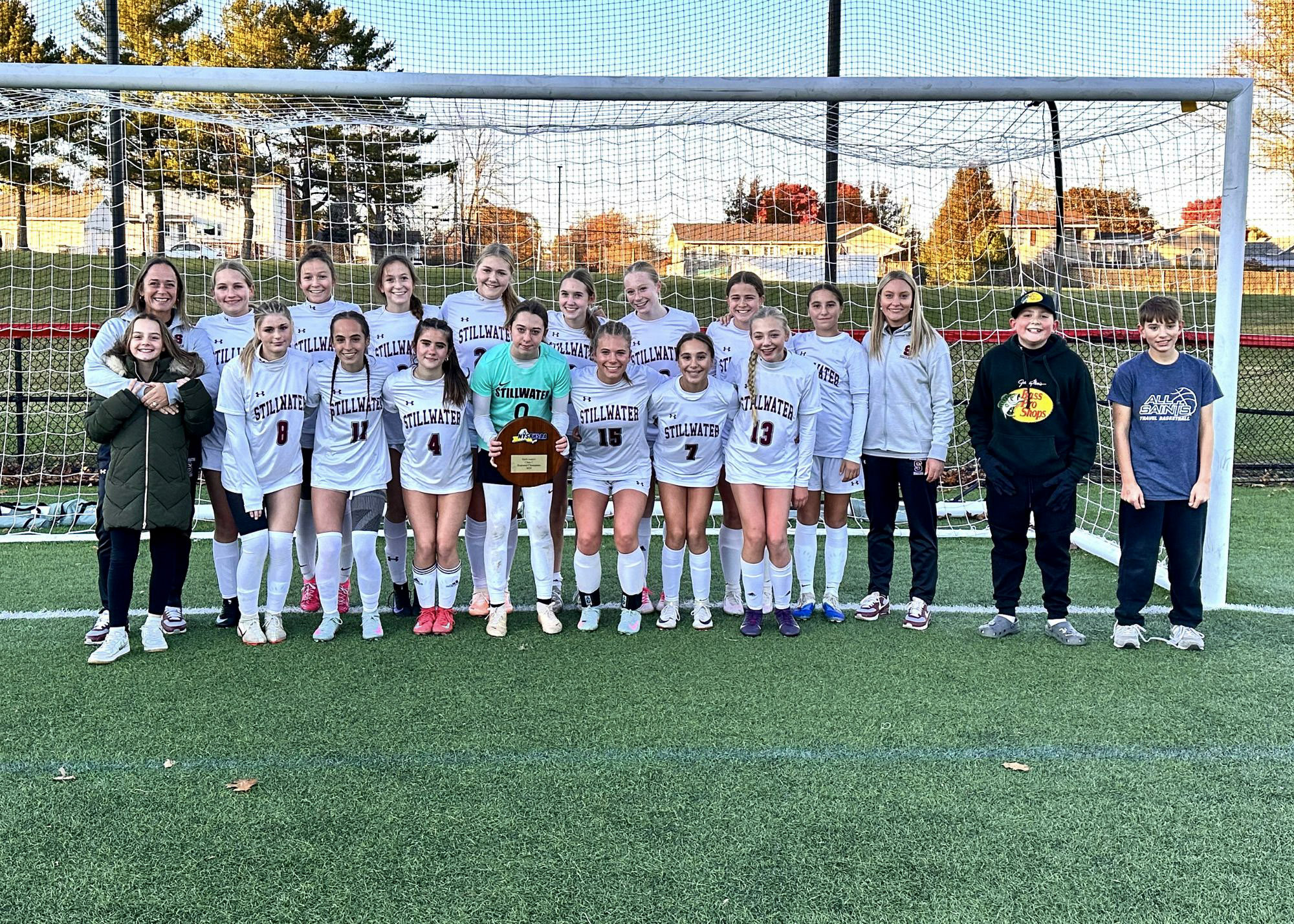 Group photo of the girls varsity soccer team in front of a soccer goal. The goalie in the middle wearing a bright green jersey is holding an award plaque.