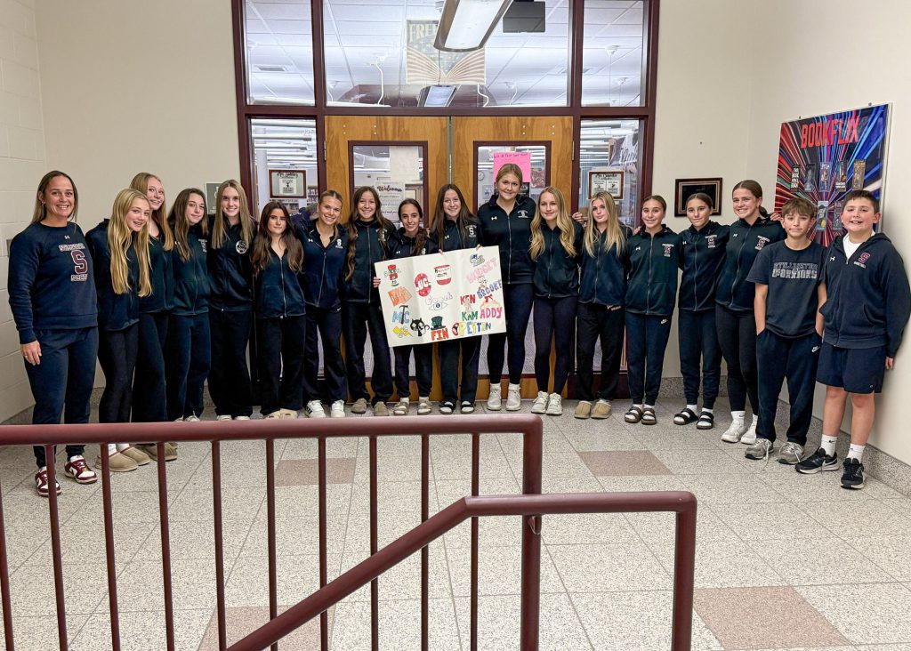 Girls varsity soccer team and their coach standing for a group photo in a school hallway, holding a sign.