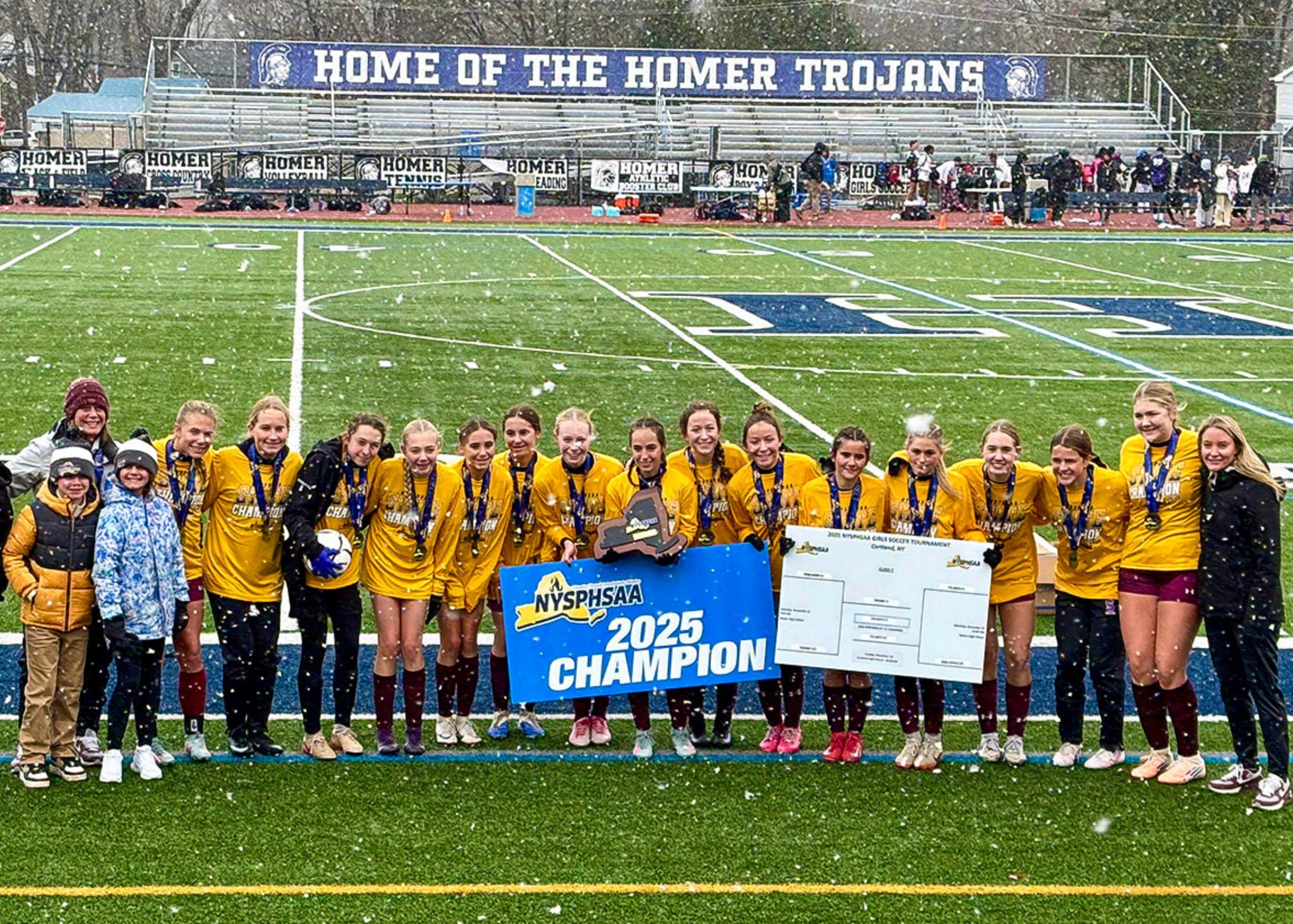 Girls varsity soccer team standing on a field outside as snow falls. They are all wearing long sleeve yellow shirts and medals around their necks. The player in the center is holding a plaque and the two players on the side of her are holding a blue banner that says "NYSPHSAA 2025 Champion."