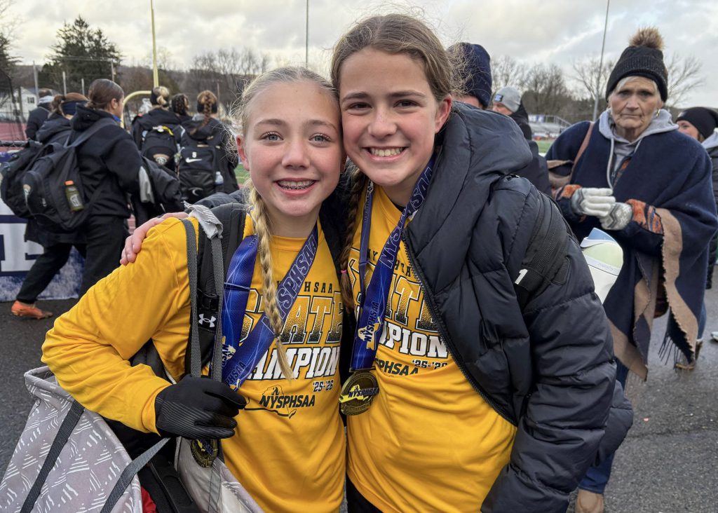 Two female soccer players wearing long sleeve yellow shirts and medals around their necks, smiling for a photo. The player on the right is wearing a heavy black winter jacket.