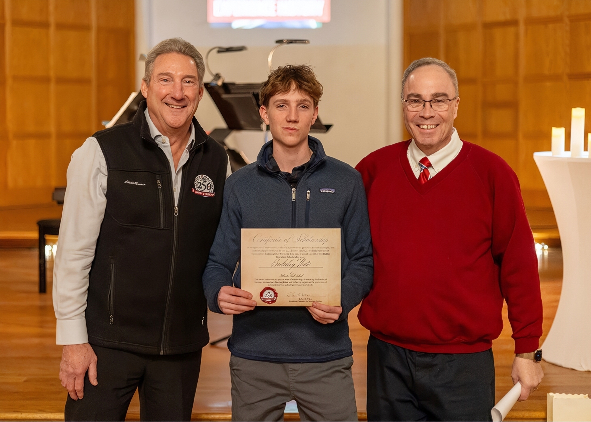 Two men and a high school student standing together for a group photo, smiling. The student is in the middle - he's wearing a dark blue Patagonia zip-up jacket and holding a certificate. The two men on the left and right have gray hair. The one on the right is wearing a red sweater with a matching tie underneath, as well as glasses. The man on the left is wearing a light colored dress shirt with a black zip-up vest.