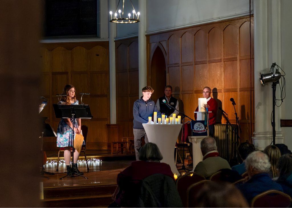 One student is speaking at a podium with the spotlight on him in a dark room. Some of the audience can be seen. There is another student on stage off to the left holding a folder. Two men also stand on stage behind the student that's speaking.