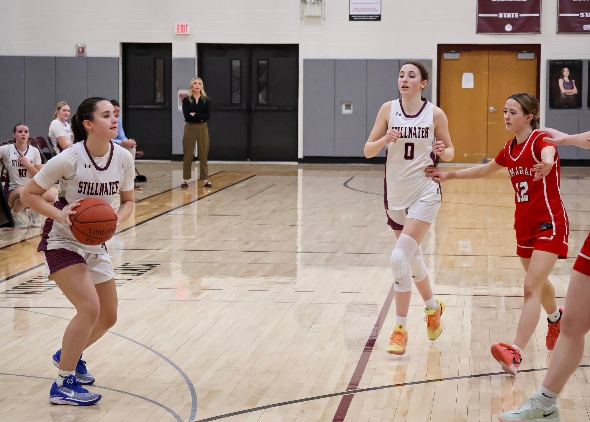 A basketball game is taking place, with a player in a white jersey dribbling the ball on the court while other players from both teams are in motion. One teammate is positioned near her, and two opponents are closely guarding her.