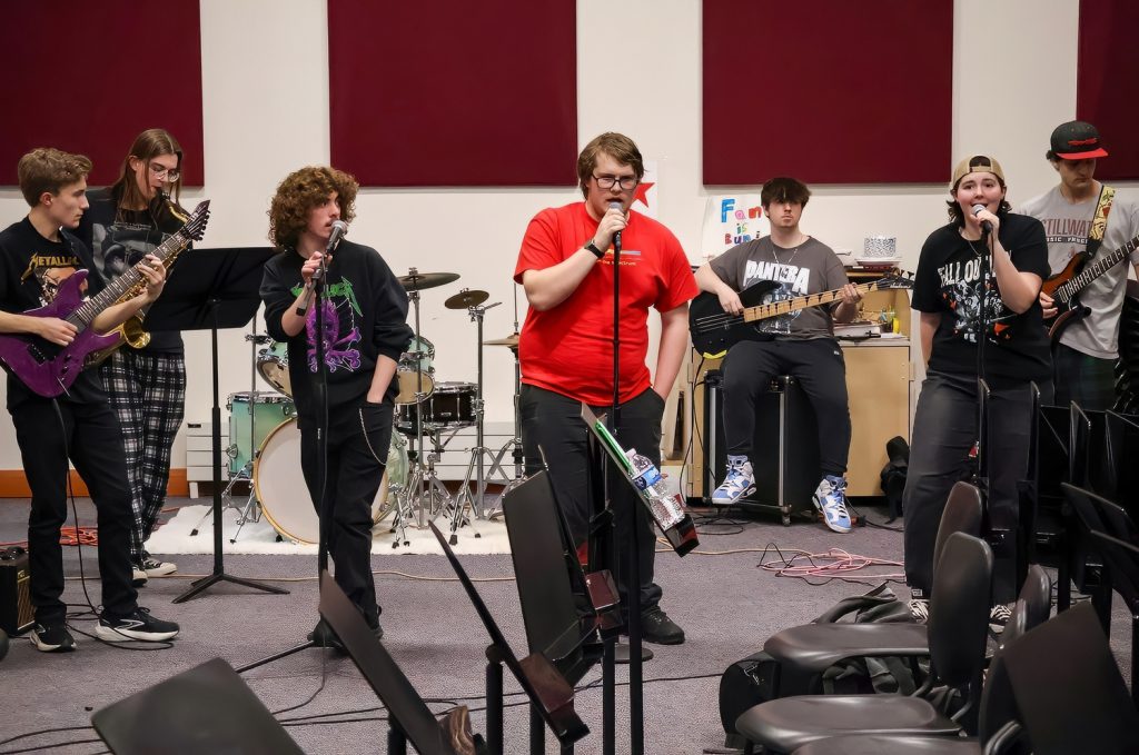 A group of seven student musicians rehearsing in a music room, with two on microphones and others playing guitars.