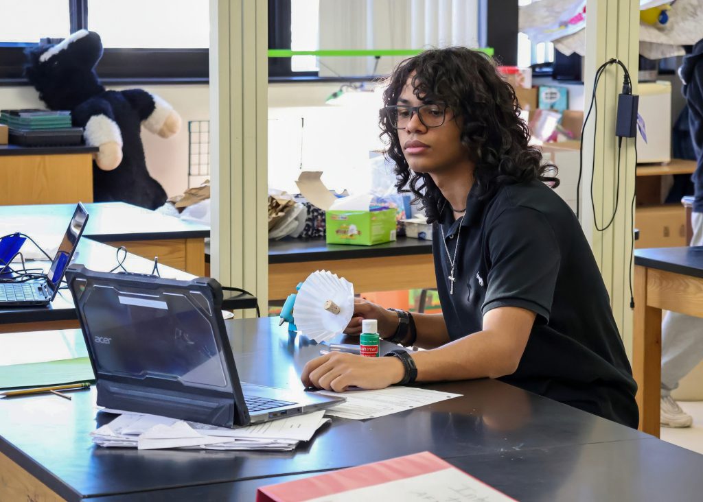 A male high school student with curly hair and glasses sitting at a table in a classroom, looking at a laptop while holding a small model. Classroom materials and tools are scattered on the table.
