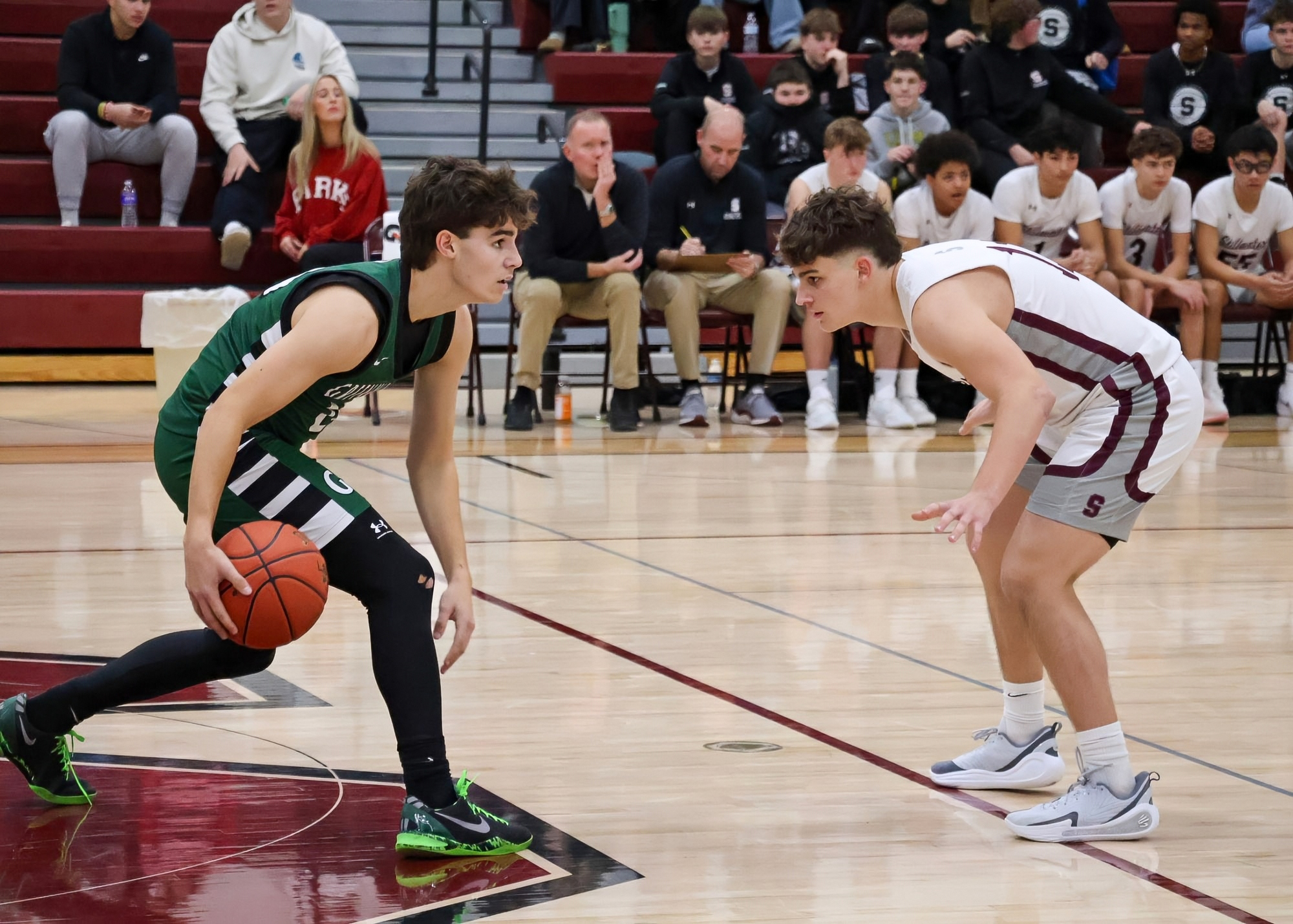 Two high school basketball players are facing each other on a court. One player is dribbling a basketball while the other is guarding him. The teams wear different colored uniforms, and there are spectators visible in the background.