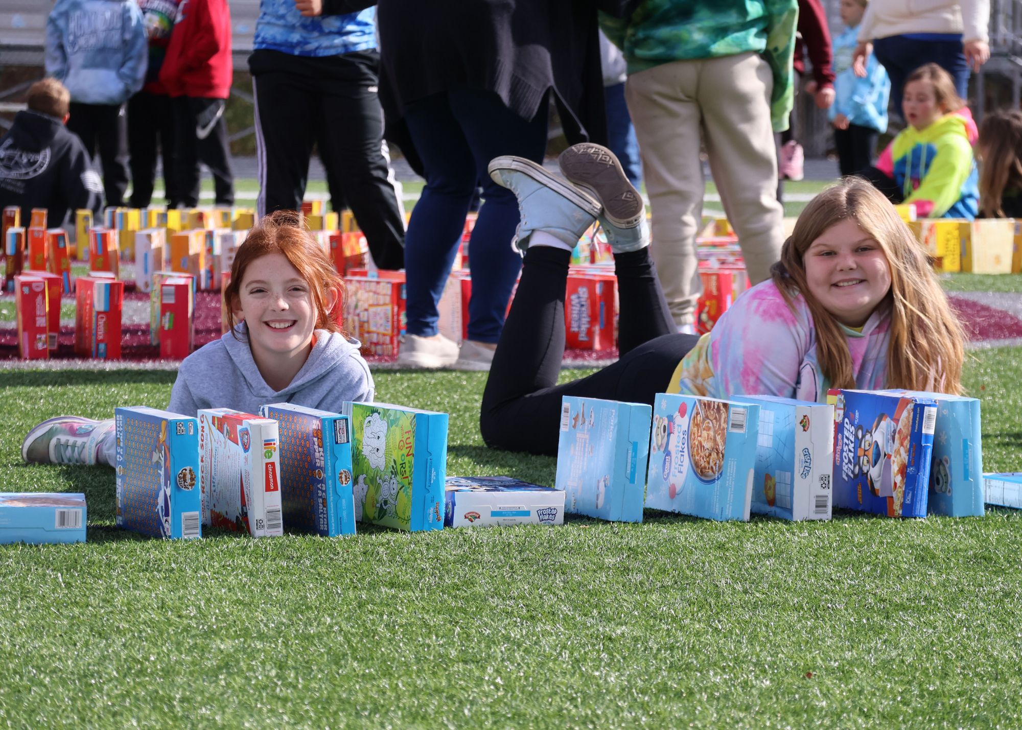 Two female elementary students lie on the turf, smiling at the camera while surrounded by boxes of cereal arranged in the background.