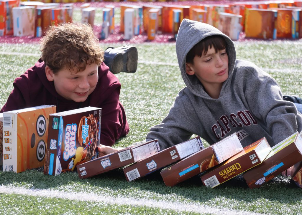 Two male elementary students lying on a turf field, trying to hold up cereal boxes that are falling down in a domino-like fashion.