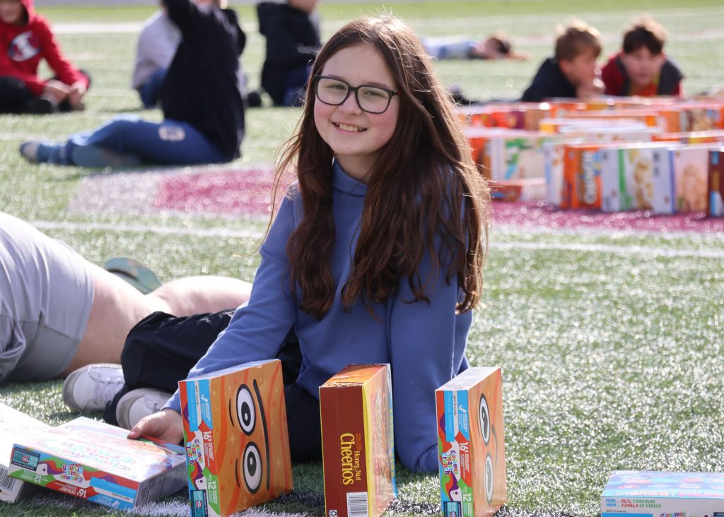 A female elementary student with glasses sitting on a turf field surrounded by boxes of cereal, smiling at the camera.