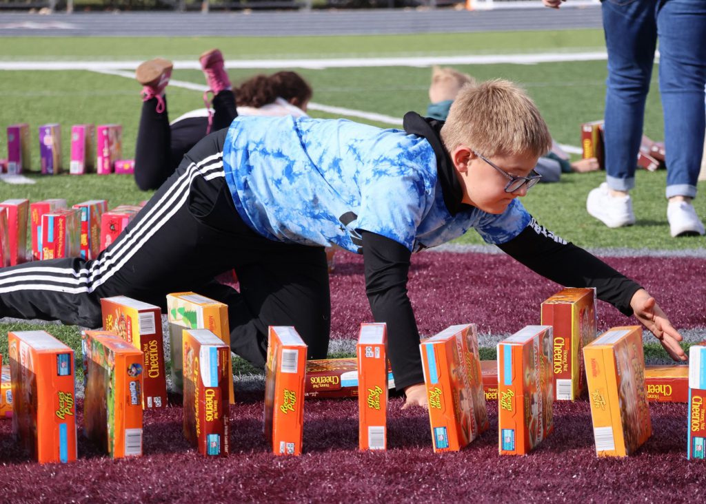 A male elementary student wearing a blue tie-dye shirt and black pants is kneeling on the ground, arranging colorful cereal boxes in a line on a turf field.