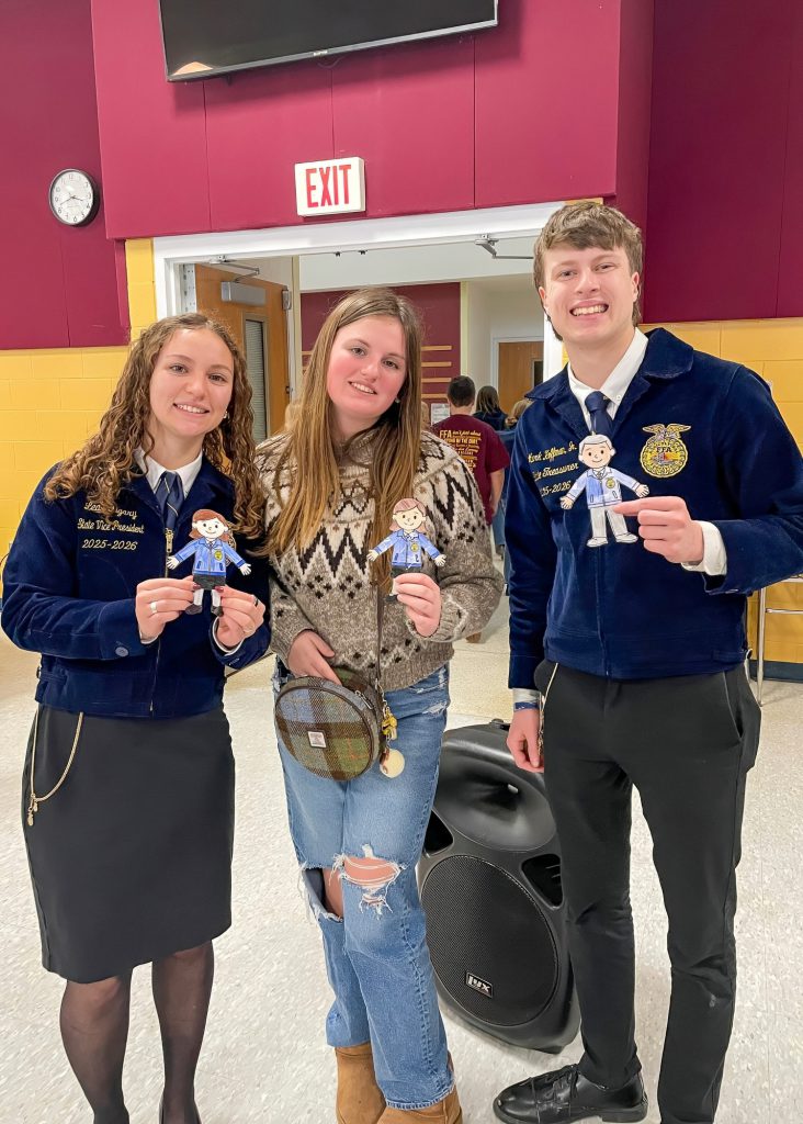 Three students in FFA jackets are holding small cut-out figures, smiling for the camera. They are standing in a school hallway, with a speaker and exit sign visible in the background.