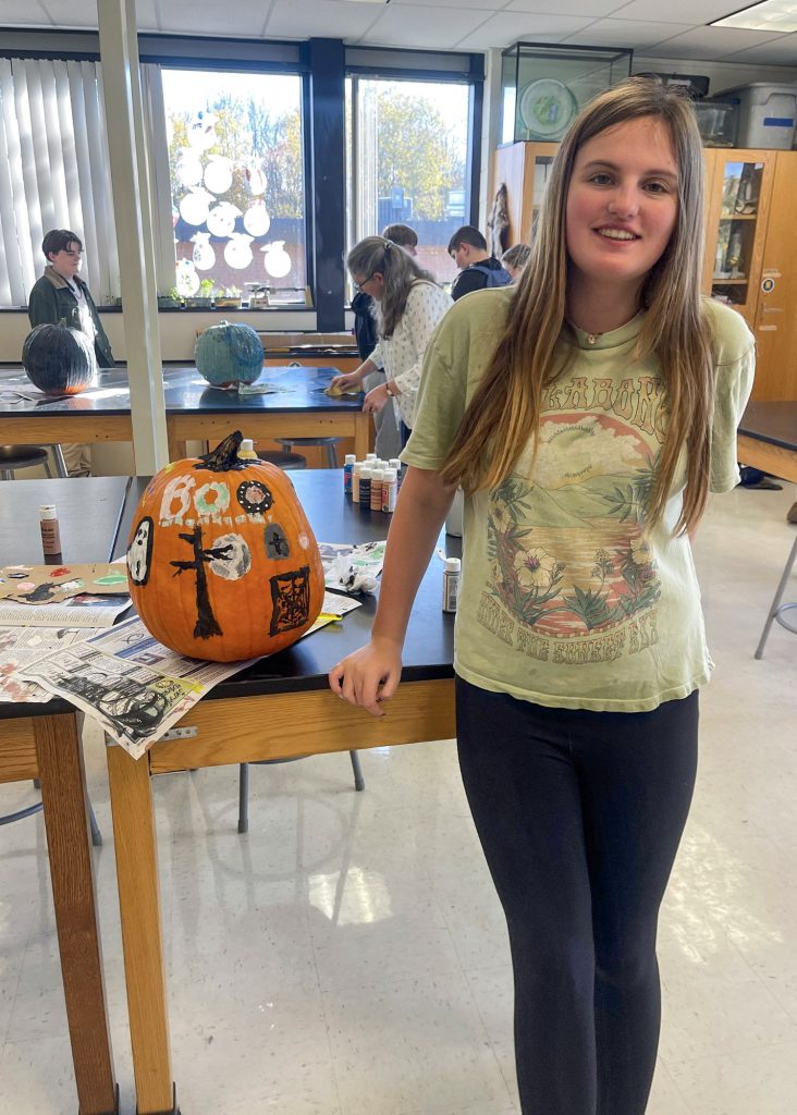 A female middle school student stands next to a decorated pumpkin on a table in a classroom, with other students working in the background.